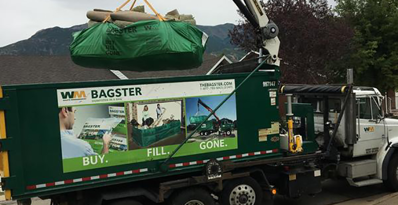 A Bagster bag filed with construction debris is loaded into a WM trash truck using a crane.